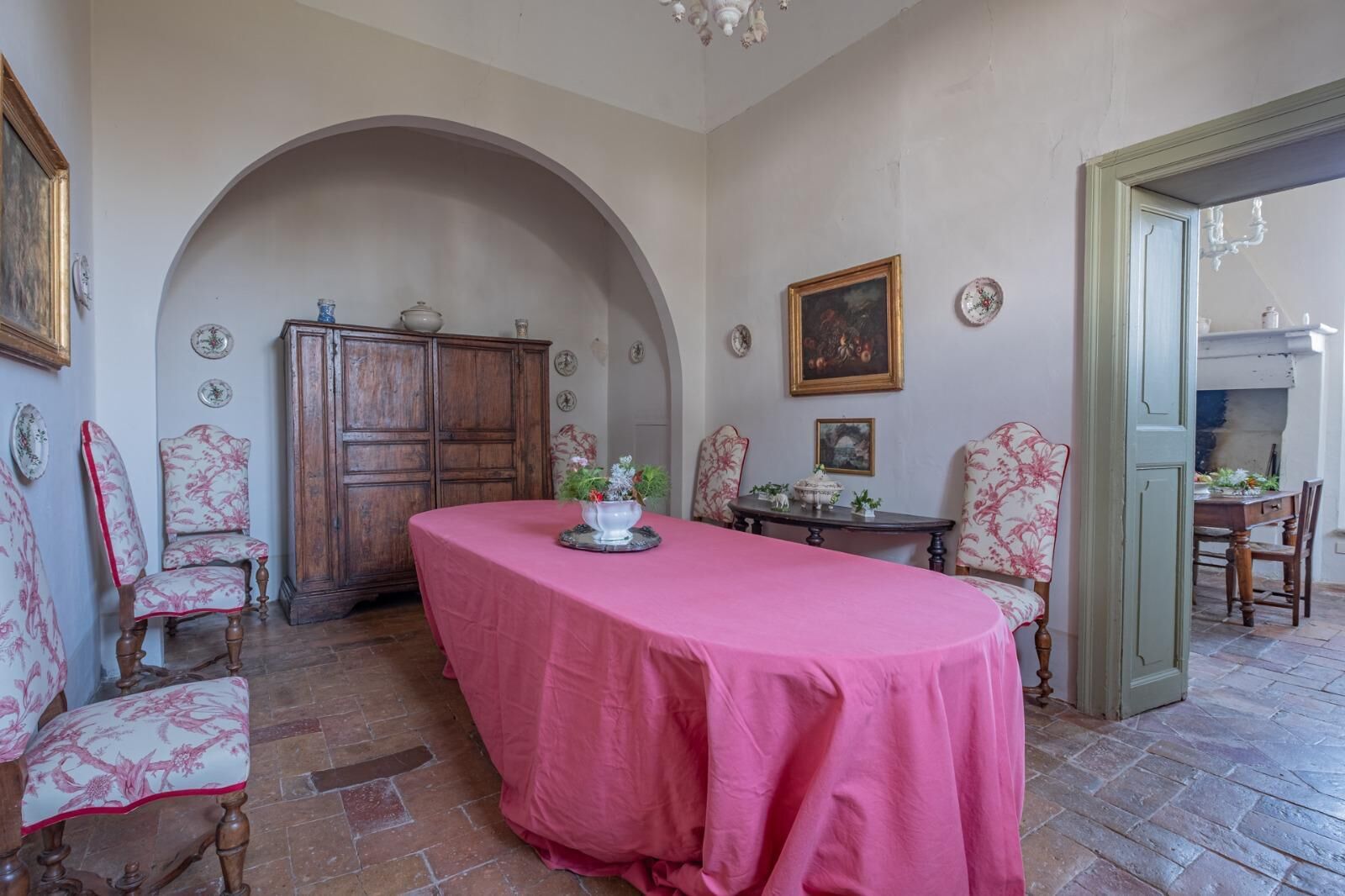A dining area with a pink tablecloth, wooden chairs, a cabinet, and a chandelier.