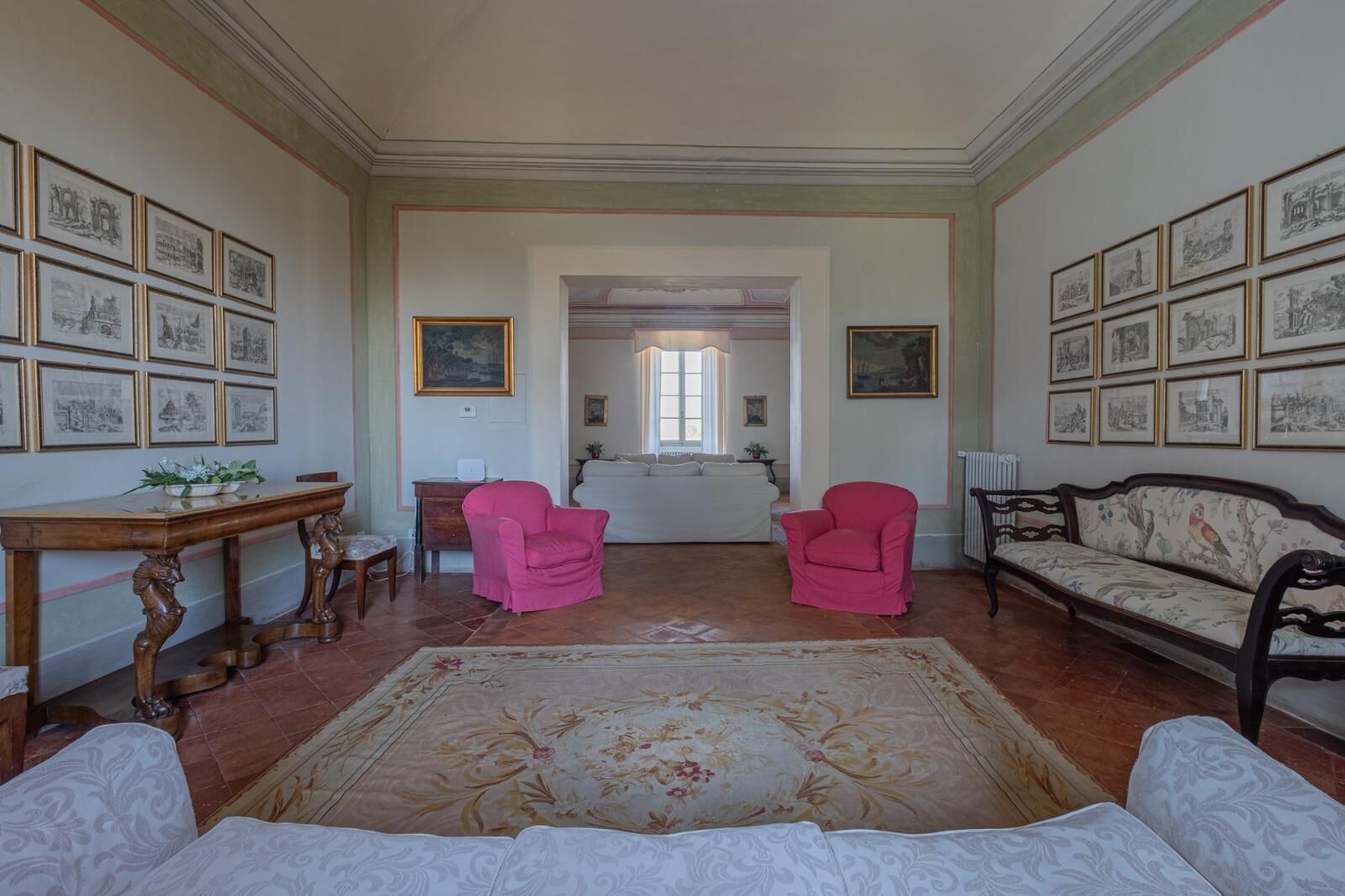 A spacious room with a large floral rug, two pink armchairs, a sofa, and a wooden table.