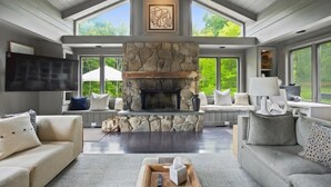 A spacious living room with a stone fireplace, large windows, and a high ceiling.