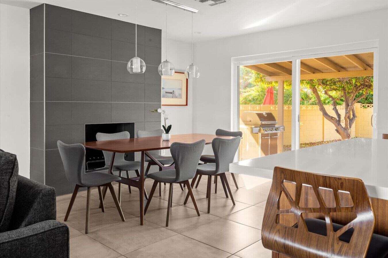 A modern dining area with a round table and six chairs, a built-in grill outside, and a large window with a view of greenery.