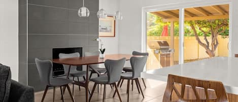A modern dining area with a round table and six chairs, a built-in grill outside, and a large window with a view of greenery.