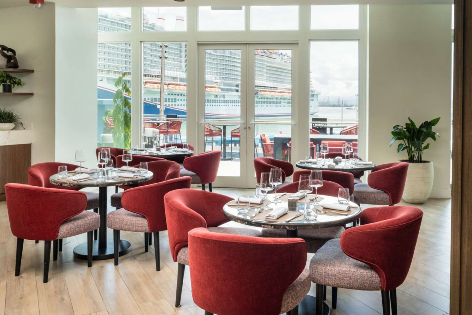 Un restaurante con mesas redondas y sillas rojas, amplias ventanas con vista a un crucero, y plantas en macetas.