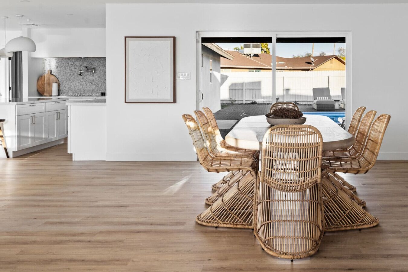 A modern dining area with a large table and wicker chairs, a kitchen with white cabinets, and a view of a residential area through a glass door.