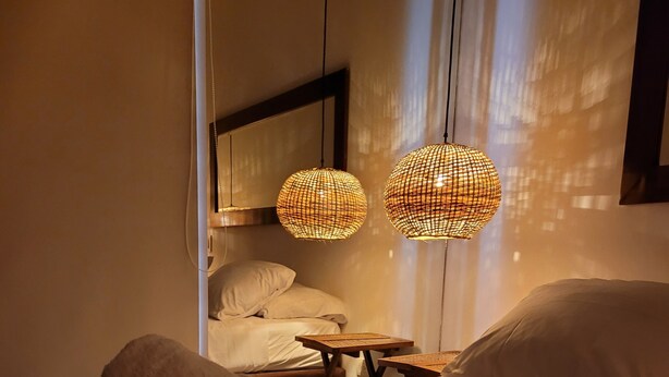 Una cama bien hecha con toallas enrolladas, una pequeña mesa de madera y dos lámparas colgantes tejidas.