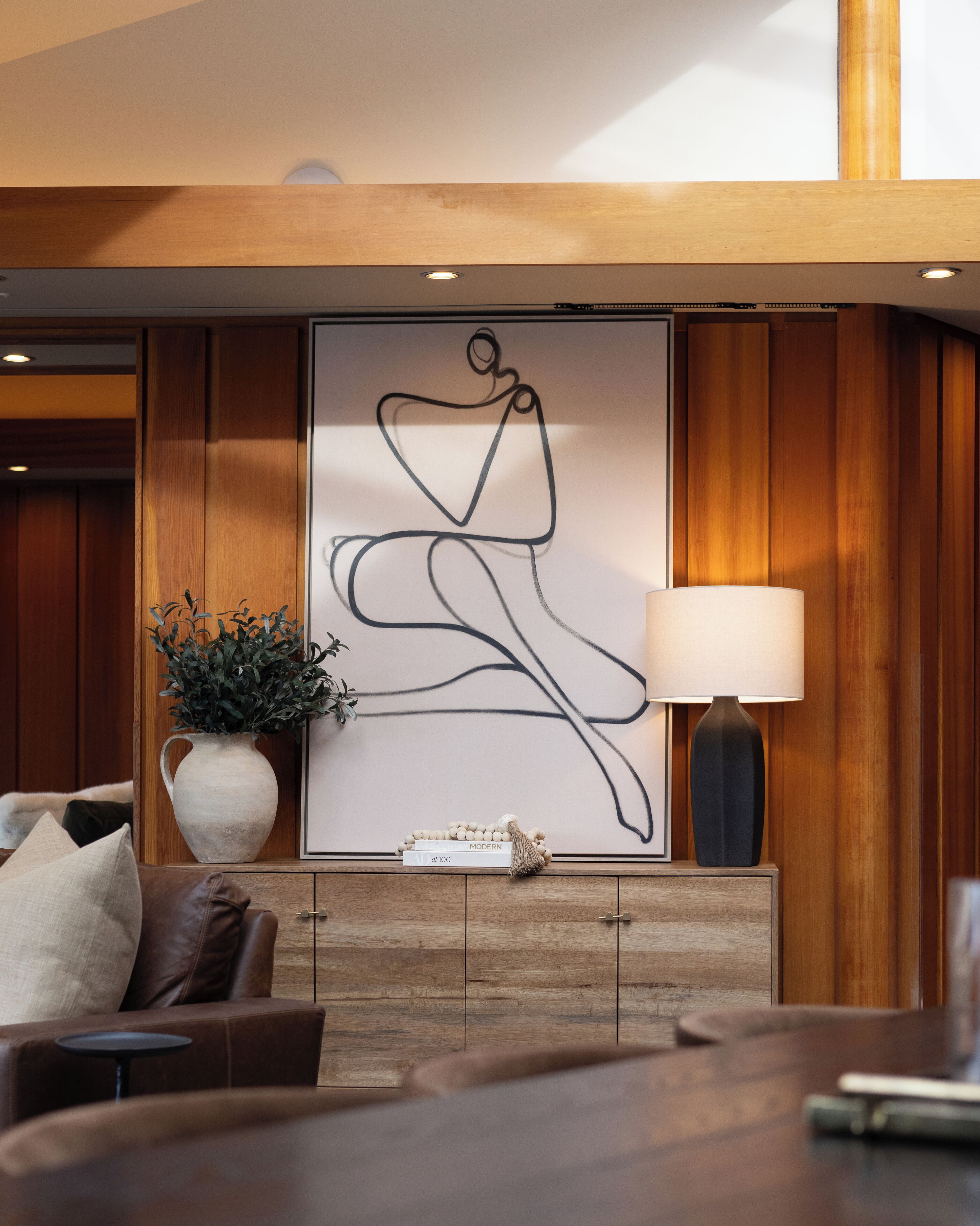 A modern living room with a wooden cabinet, a large abstract painting, a potted plant, and a lamp.