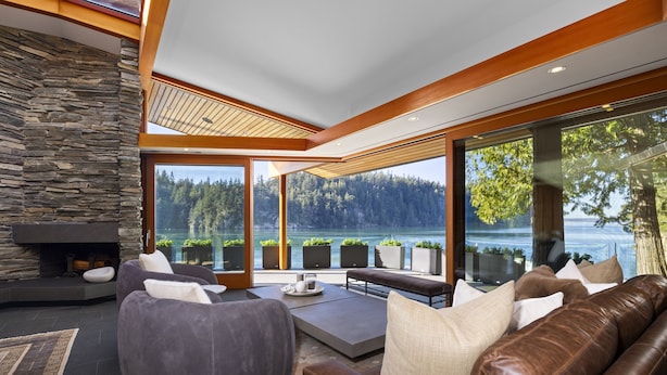 Una sala de estar moderna con chimenea de piedra, ventanas amplias y vistas a un lago y bosque.
