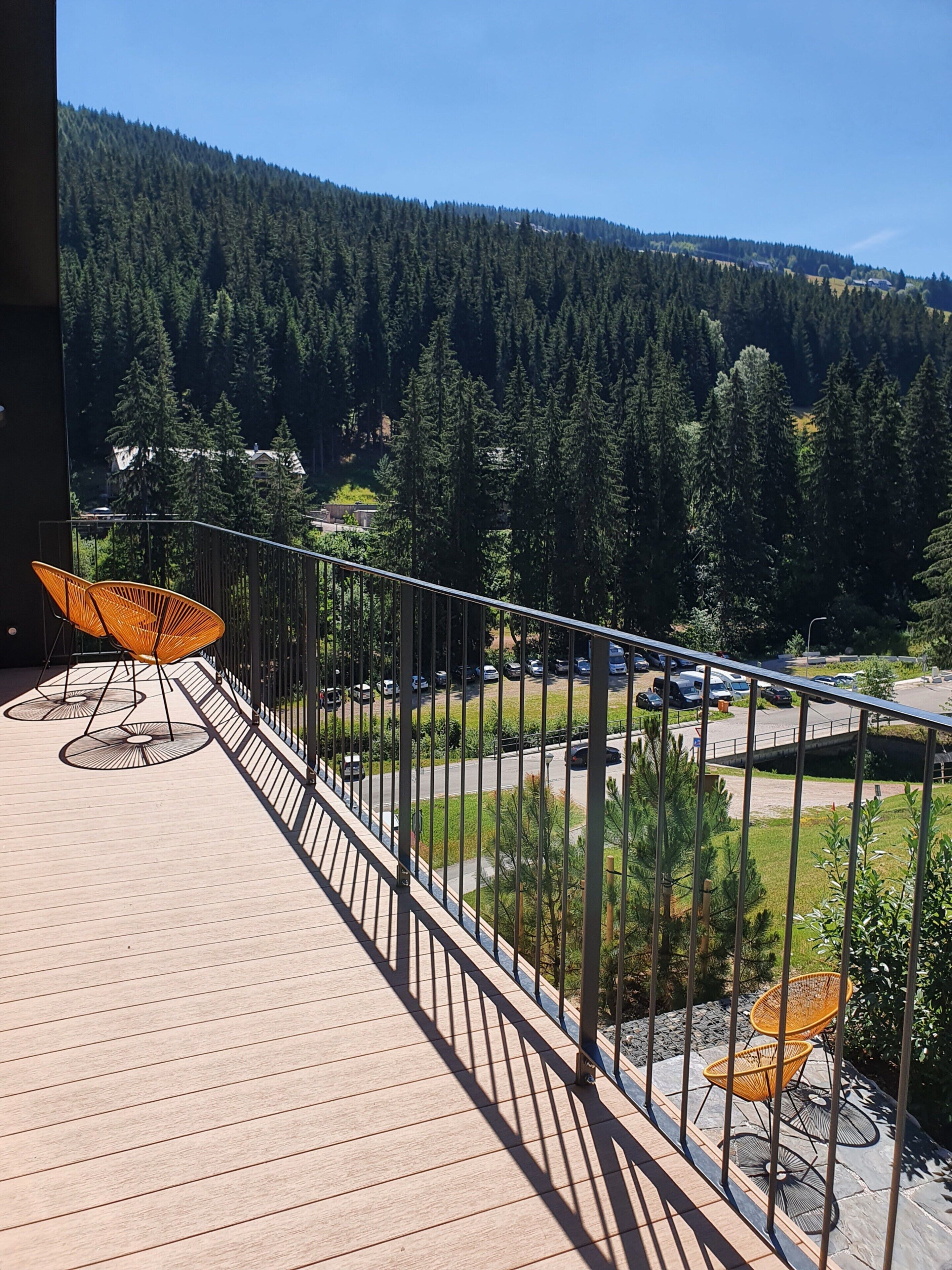 A wooden deck with outdoor furniture, a metal railing, and a view of a forested area with a parking lot.