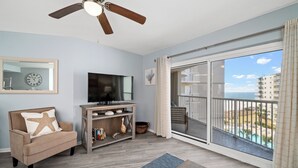 A living room with a TV, a starfish pillow, a wooden console, and a view of a balcony with a pool. - ☀Sundazed☀ ~ Spectacular Beachside Condo w/Fabulous Pool at Tradewinds in OBA⛱ (Orange Beach)