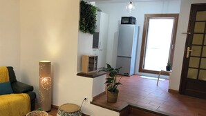 Ein Wohnzimmer mit Sofa, einer Lampe, einem kleinen Tisch und einem Blumentopf. Es gibt eine Küchenzeile mit Kühlschrank und Mikrowelle.