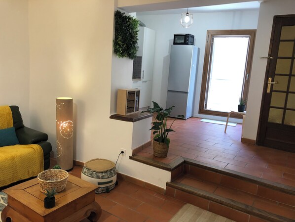 Ein Wohnzimmer mit Sofa, einer Lampe, einem kleinen Tisch und einem Blumentopf. Es gibt eine Küchenzeile mit Kühlschrank und Mikrowelle.
