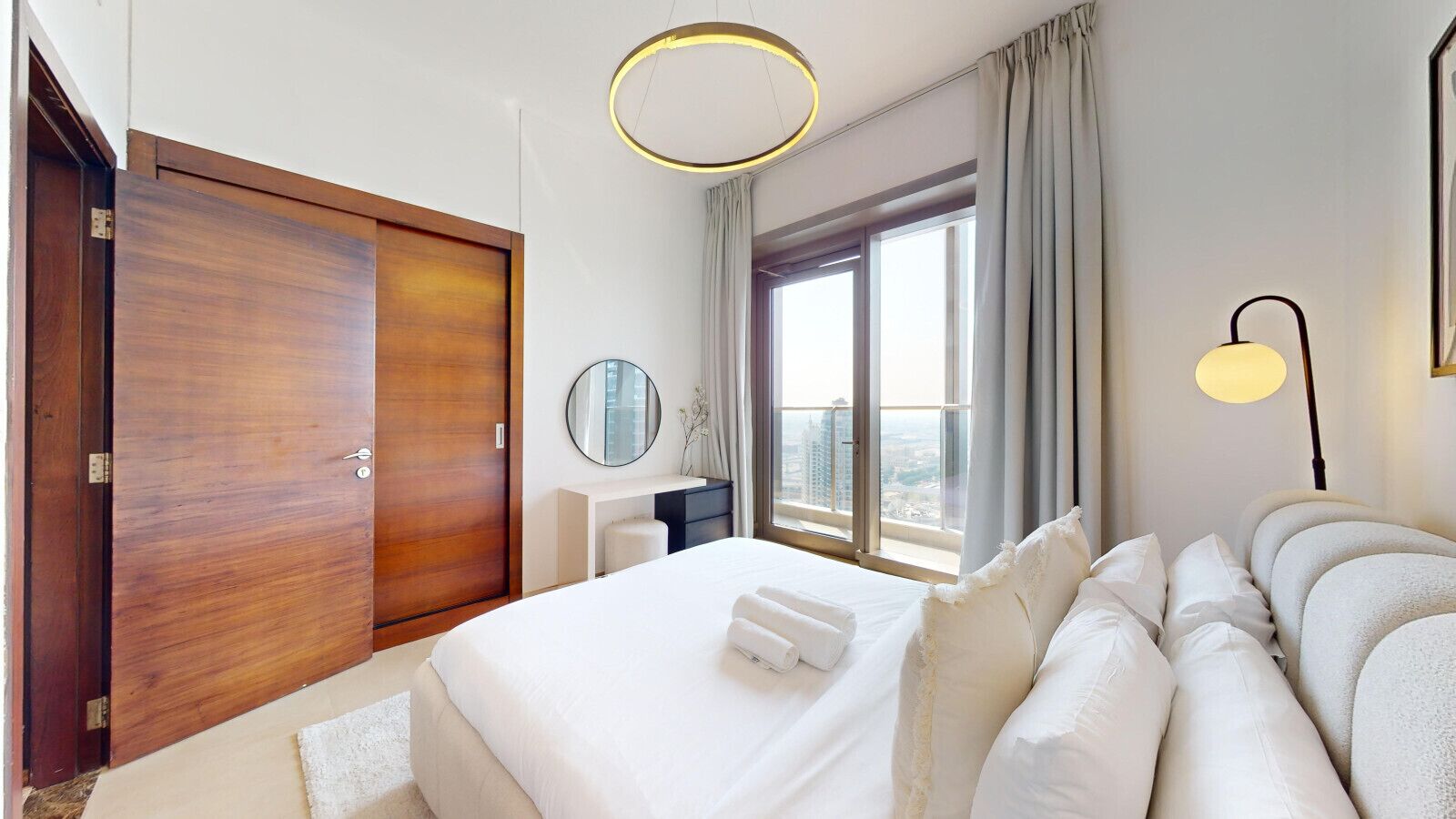 Ein Hotelzimmer mit einem großen Bett, einem Holzschrank, einem runden Spiegel, einem Nachttisch, einer Lampe und einem Fenster mit Blick auf Gebäude.