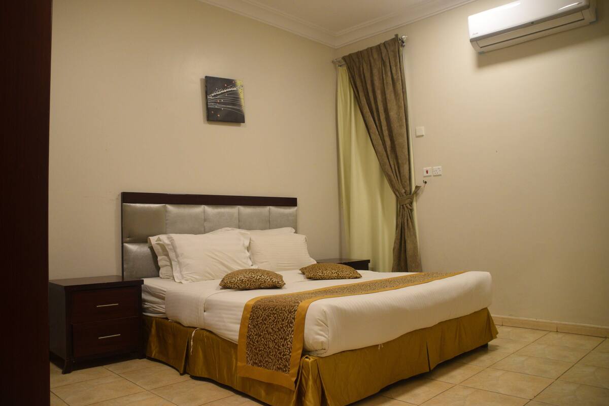 A neatly made bed with a headboard, a bedside table, and a wall-mounted air conditioner.
