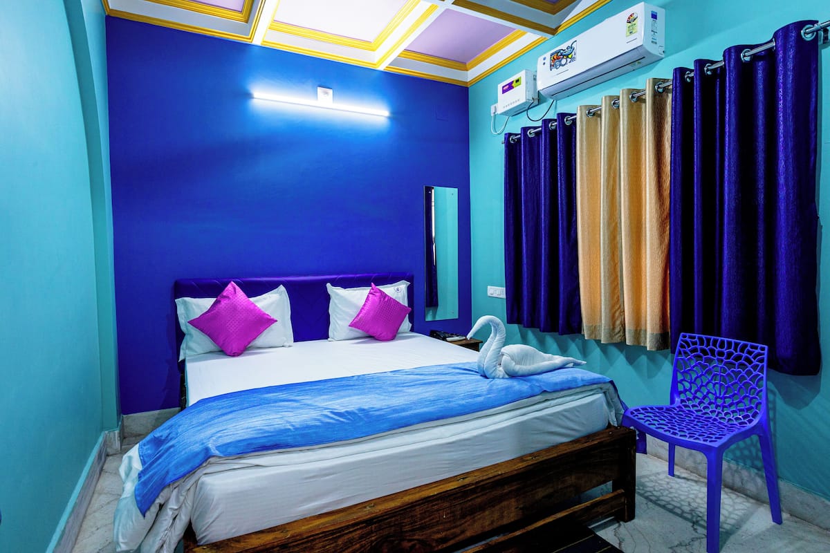 Quarto com cama, cabeceira de madeira, uma cadeira roxa e janela com cortinas.
