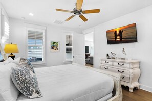 Une chambre à coucher avec un lit, un ventilateur de plafond, une télévision, une commode et un tableau représentant deux personnes sur une plage.