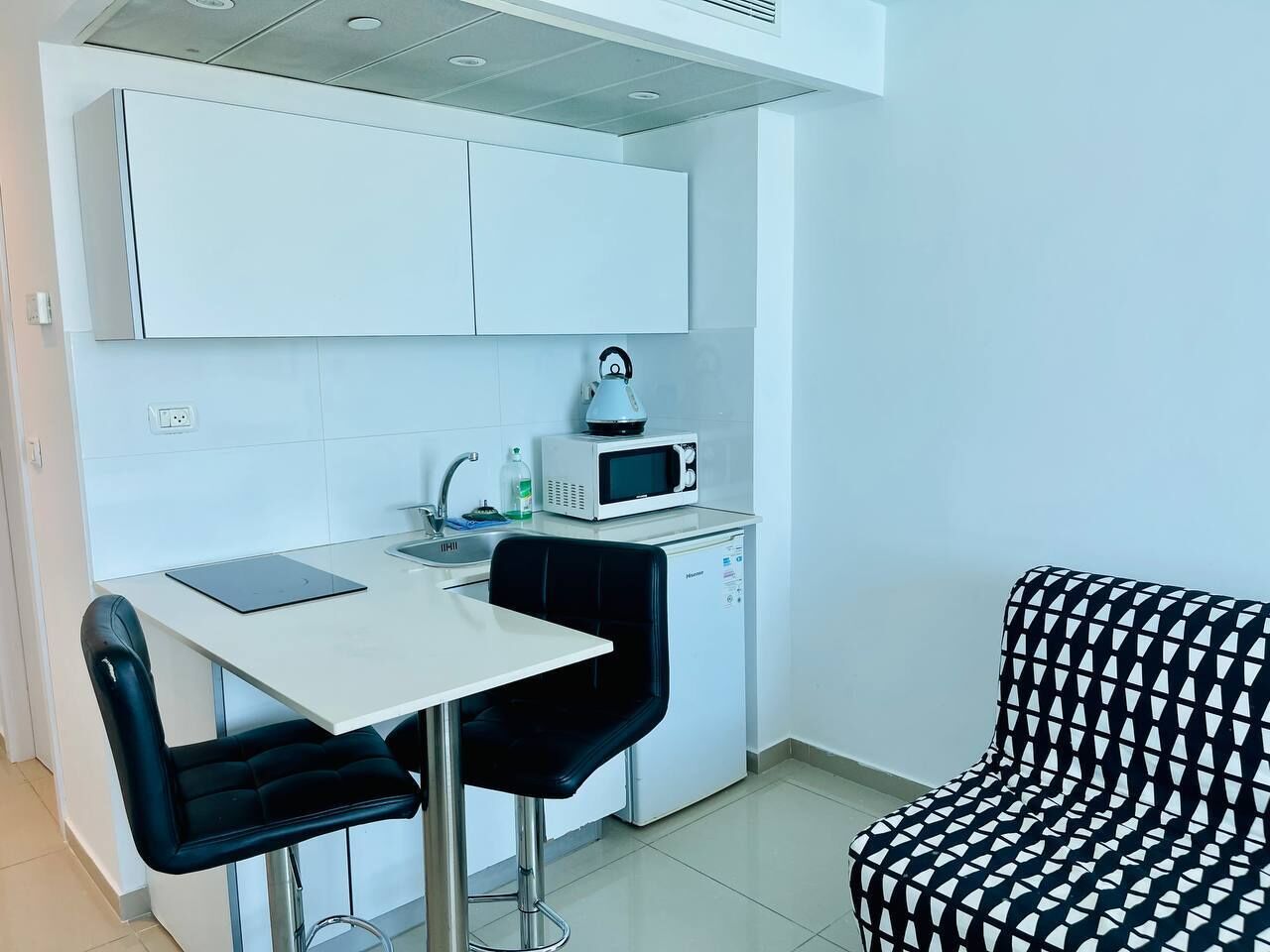 Cocina compacta con armarios blancos, un microondas, hervidor de agua y un refrigerador pequeño. Hay una mesa de comedor con una silla y un sofá estampado.