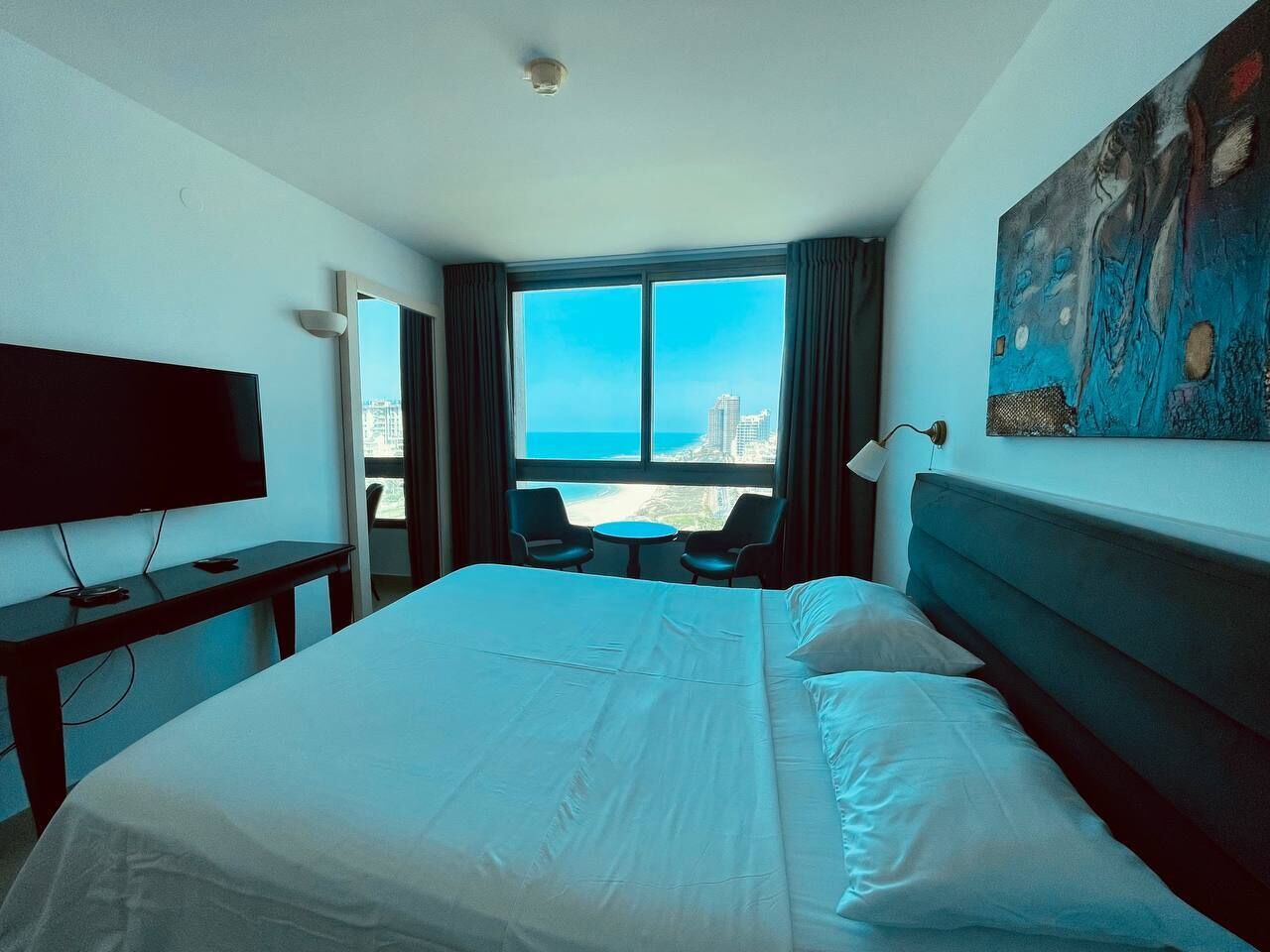 Quarto de hotel com uma cama grande, televisão, escrivaninha e vista para a cidade e oceano.