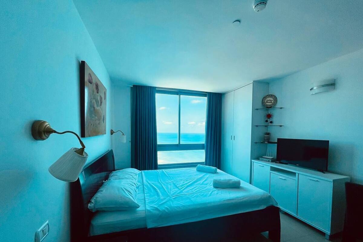 A bedroom with a bed, a TV, a desk, and a window with curtains.