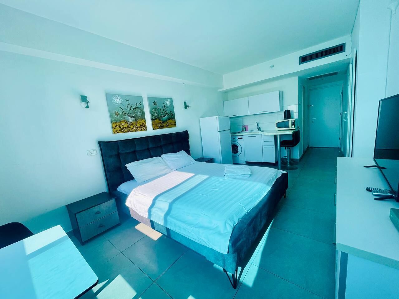 A modern hotel room with a bed, bedside table, desk, and a kitchenette.