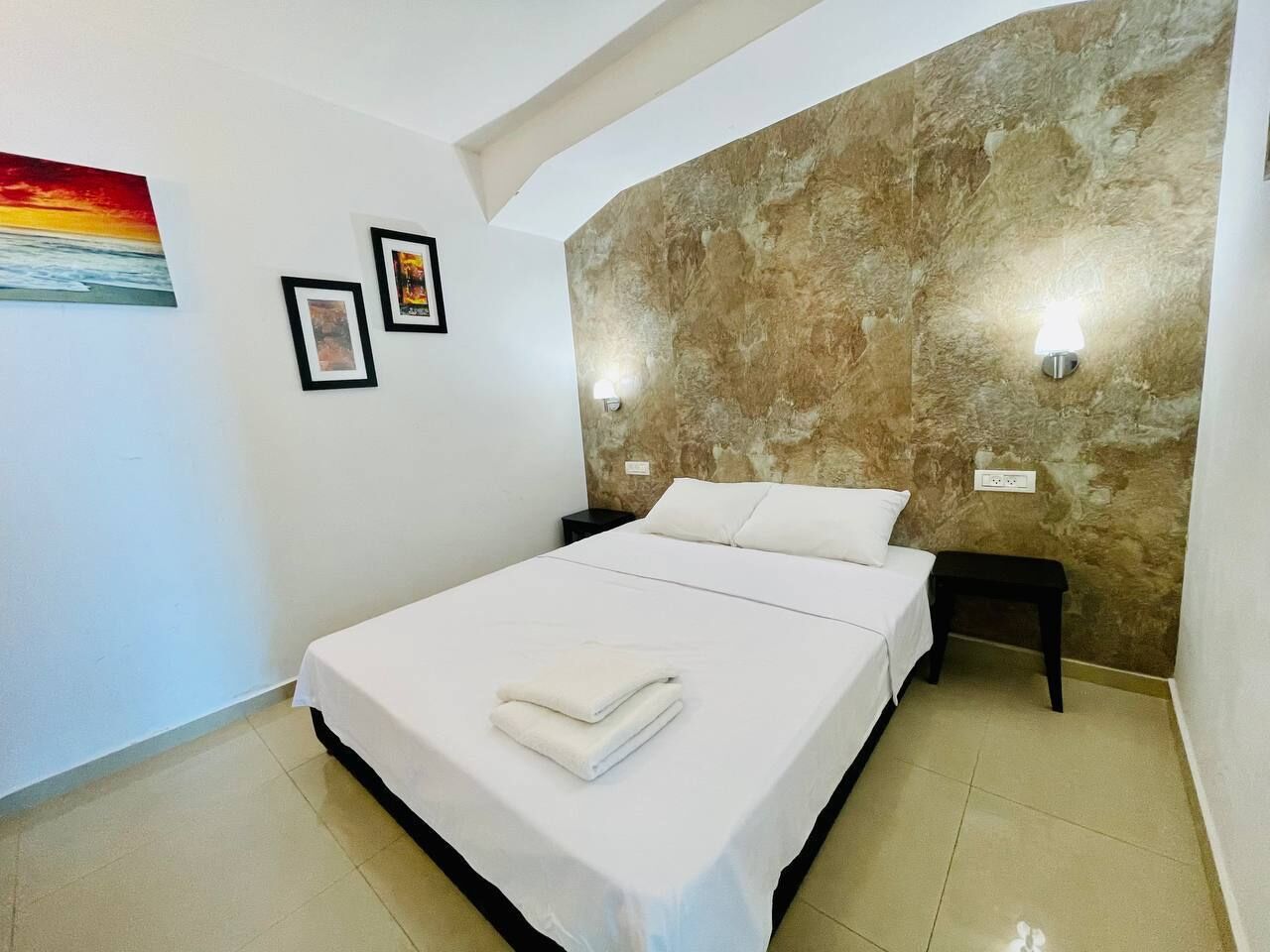 A neatly made bed with white linens and a folded towel, a wall with a textured stone-like finish, and two framed pictures on the wall.