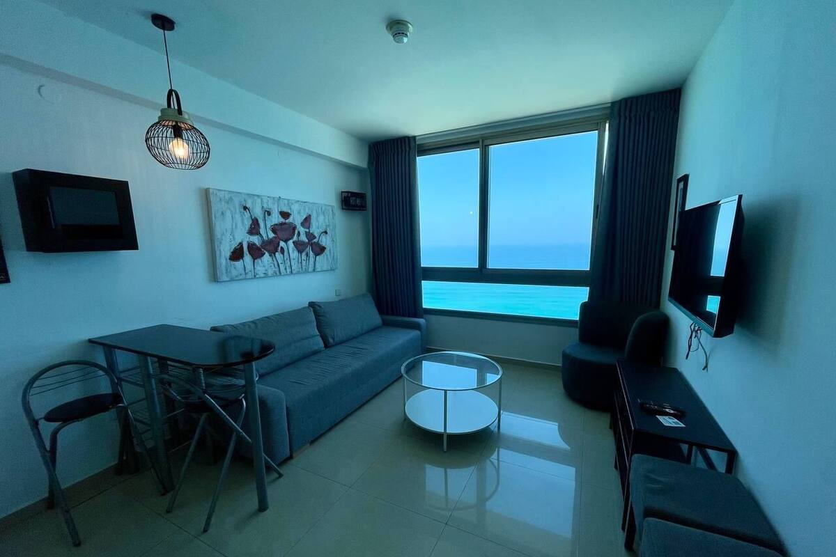 A modern living room with a sofa, a coffee table, a TV, and a large window offering a view of the sea.