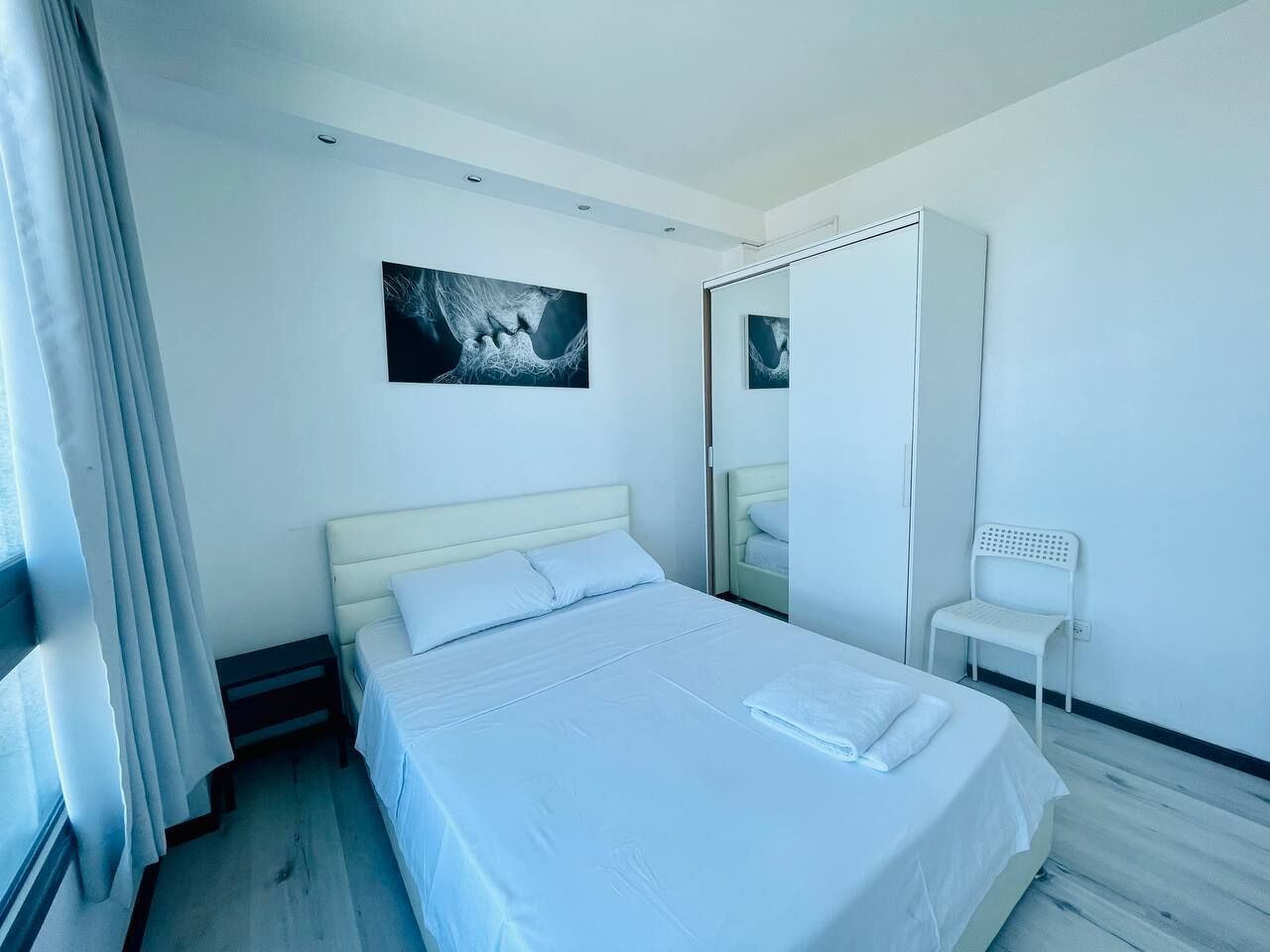 A modern hotel room with a large bed, a bedside table, a chair, a wardrobe, and a painting on the wall.