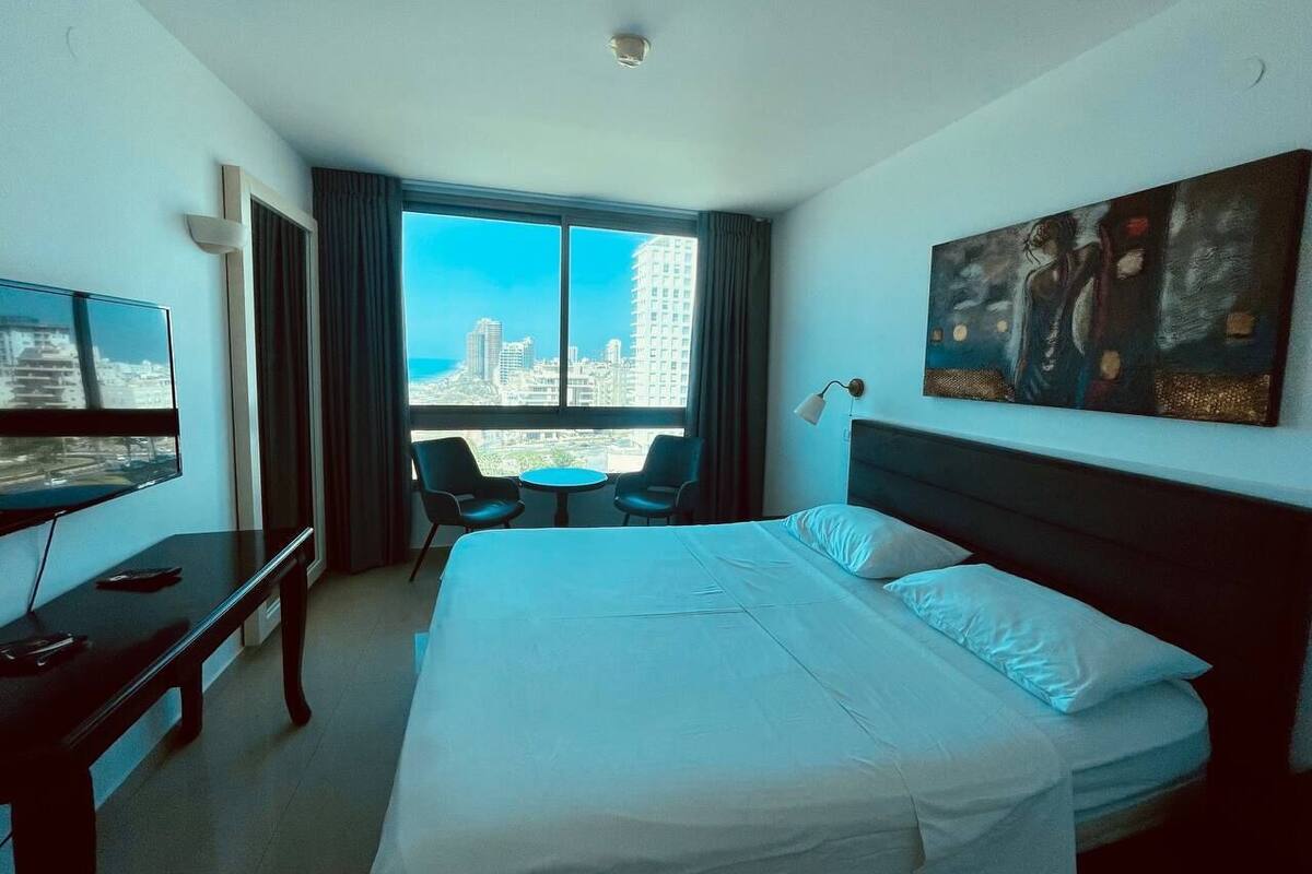 A hotel room with a large bed, a desk with a TV, a chair, a small table, and a painting on the wall.