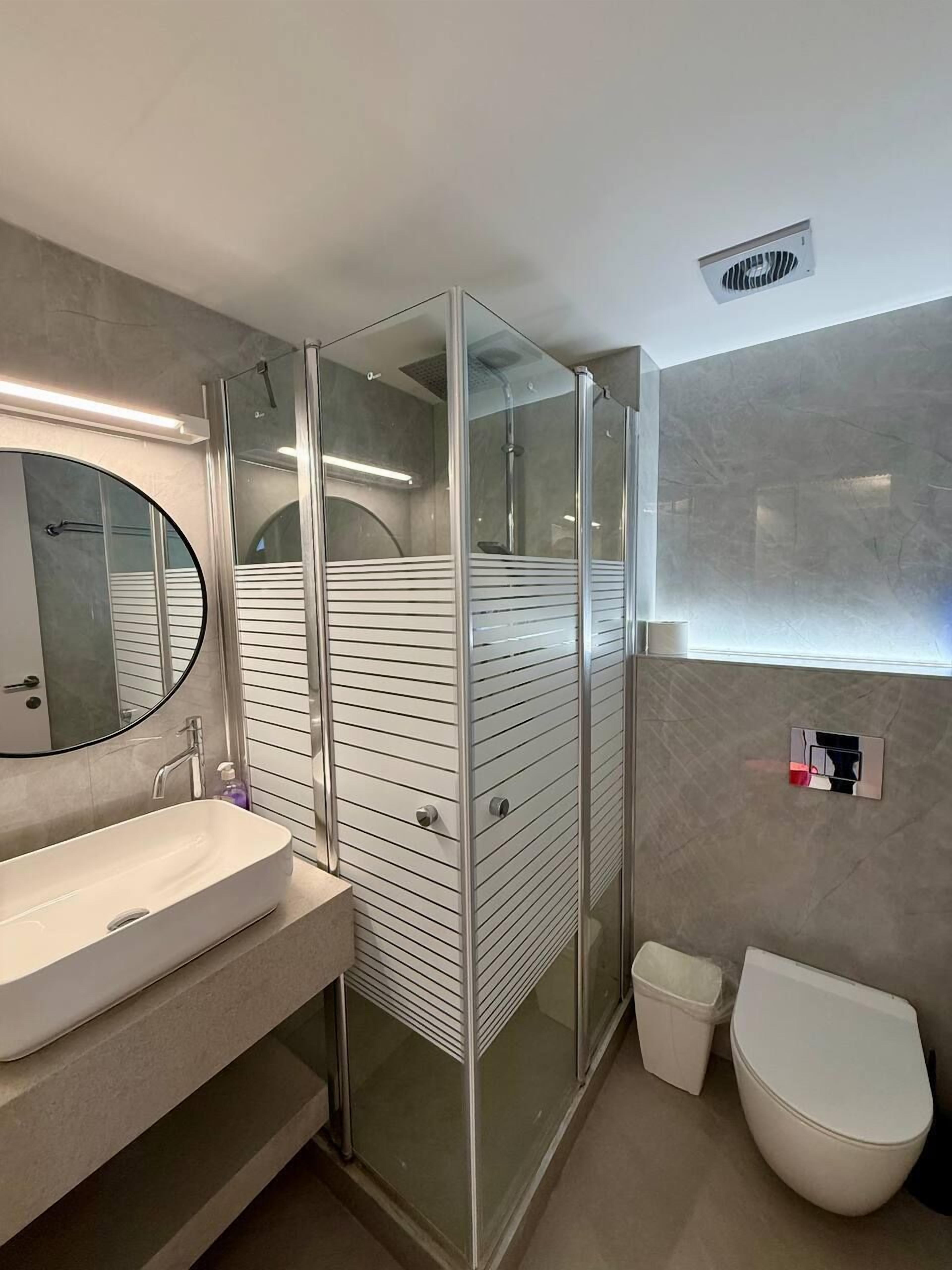 Un baño moderno con ducha de vidrio, un espejo redondo, un lavabo blanco y un inodoro.