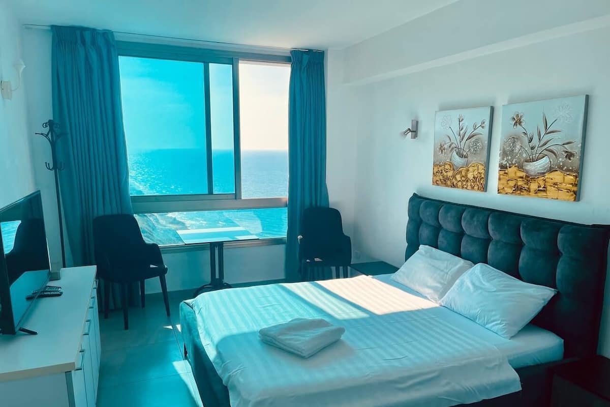 A hotel room with a bed, a desk, a chair, and a view of the sea.