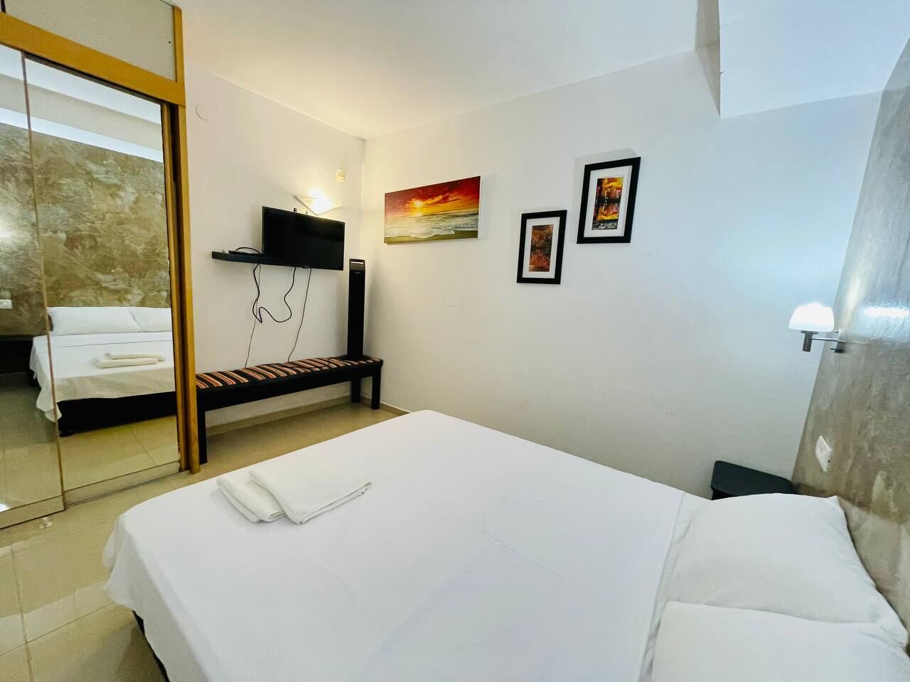 Una habitación de hotel con cama, un banco, un televisor y obras de arte en la pared.