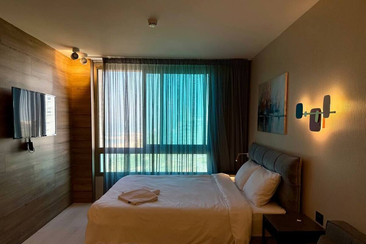 A modern hotel room with a large bed, a flat-screen TV, a painting on the wall, and a window with sheer curtains.