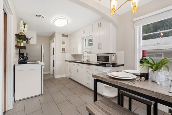 A modern kitchen with white cabinets, a black countertop, and a dining area with a wooden table and bench. - The Rams' Den – King Beds, Fire Pit, Spacious Yard (Fort Collins)