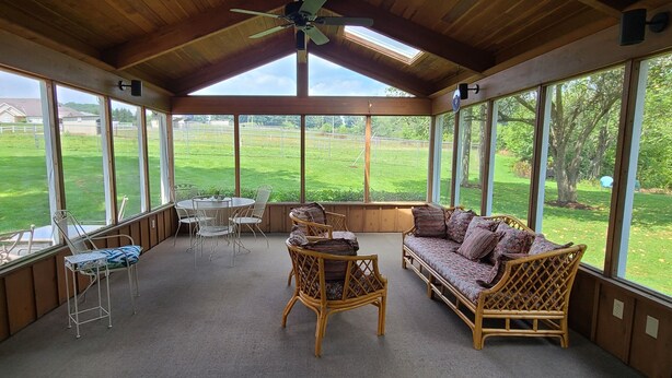A covered patio with a wooden ceiling, large windows, a round table with chairs, a sofa, and wicker furniture.