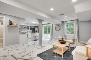 A spacious living area with a marble-patterned floor, a kitchen in the background, and a large window with curtains. - DMV 6BR Lakeside Luxury Retreat w/ Firepit, Gameroom, Pool-table (Gaithersburg)
