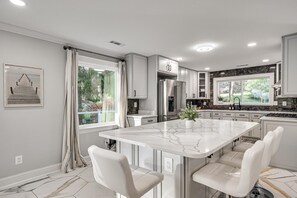 A modern kitchen with a marble island, white cabinetry, and a large window overlooking a garden. - DMV 6BR Lakeside Luxury Retreat w/ Firepit, Gameroom, Pool-table (Gaithersburg)