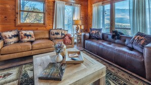 A cozy cabin interior with two leather sofas, a wooden coffee table, and a view of the mountains through large windows. - Majestic Cabin Getaway • Epic Views, Hot Tub & Game Room • Family-Friendly (Gatlinburg)