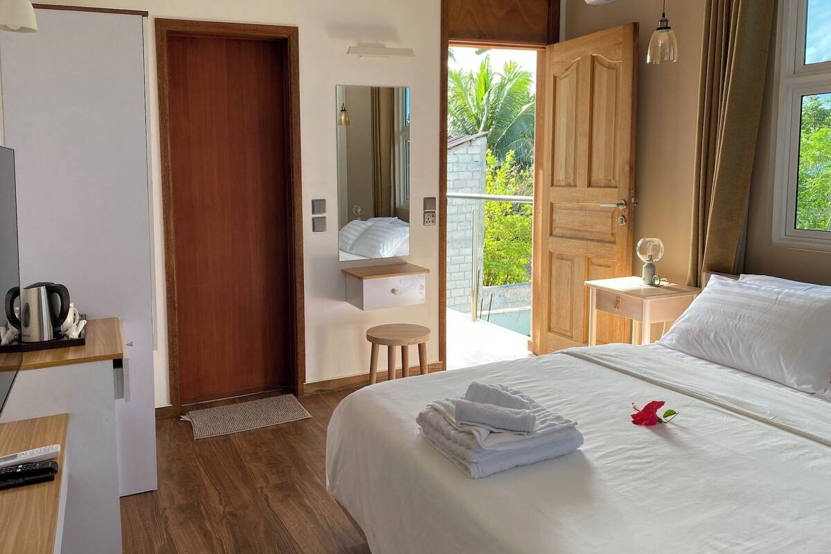 A hotel room with a bed, a wooden desk, a small stool, a mirror, a door leading to a balcony, and a window with curtains.