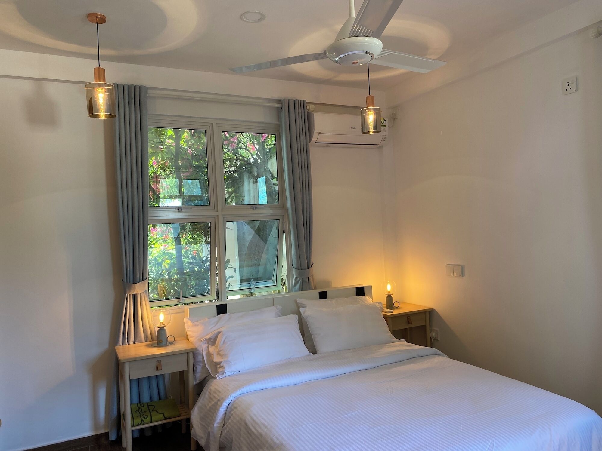 Une chambre à coucher comprenant un lit, deux tables de chevet, une commode, un ventilateur de plafond et une fenêtre avec des rideaux.