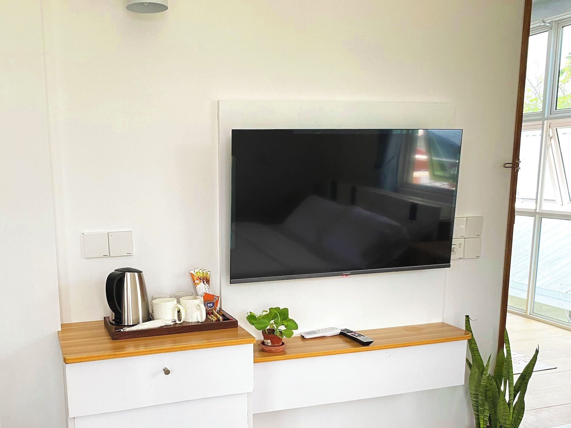 A modern hotel room with a flat-screen TV mounted on the wall, a wooden side table with a kettle, cups, and a plant, and a large window offering a view of the outdoors.