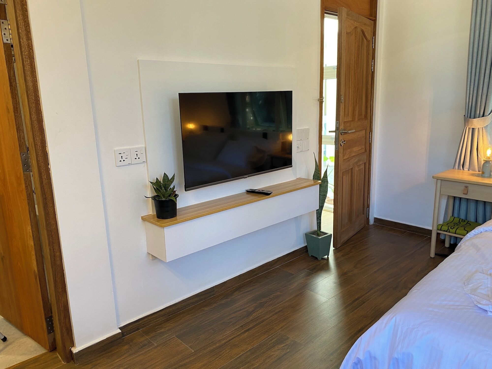 Un dormitorio moderno con una televisión de pantalla plana montada en una pared blanca, suelo de madera, una mesita de noche con lámpara y una pequeña planta en maceta.