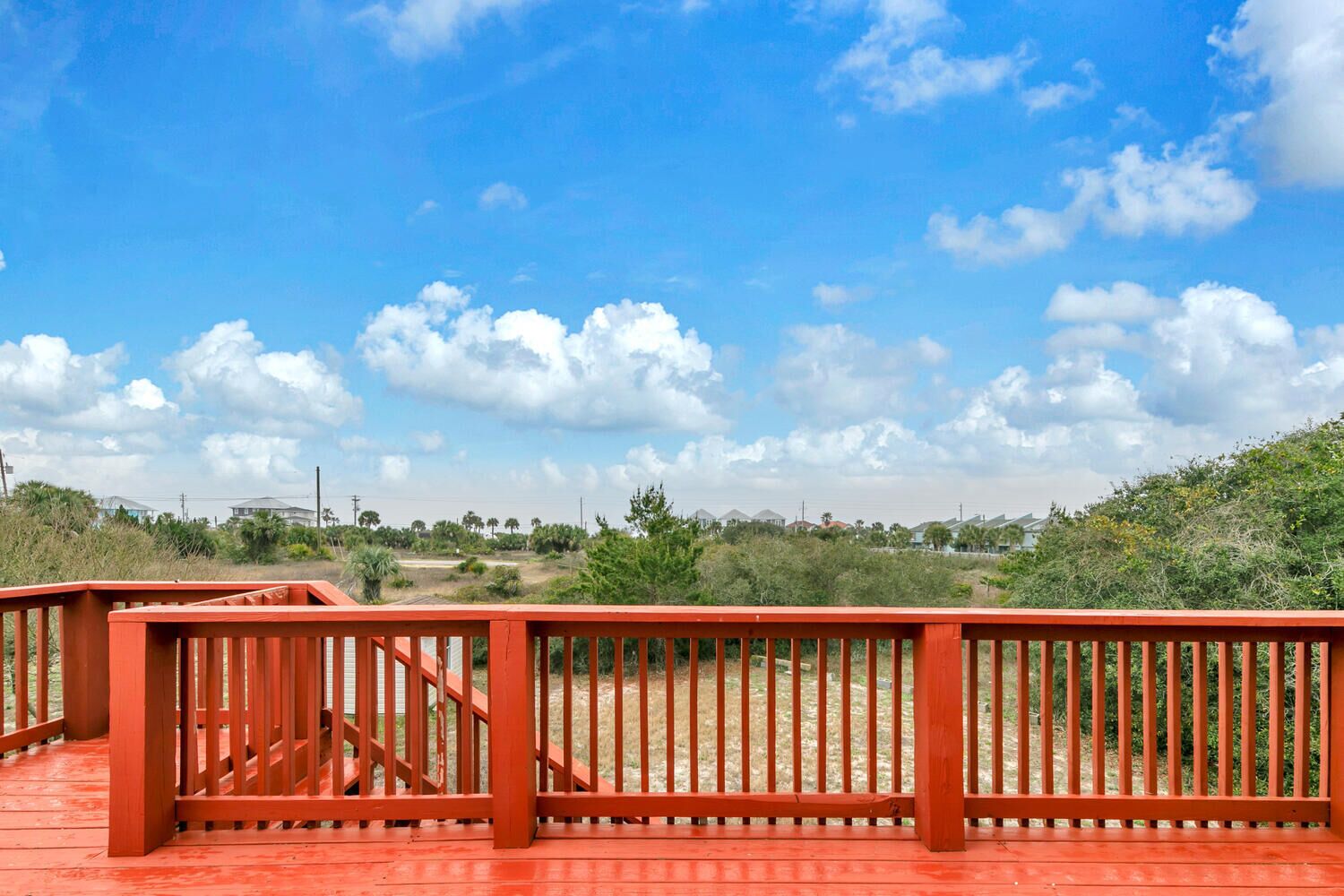 Een rood houten terras met een hekwerk, met uitzicht op een landschap met huizen en begroeiing onder een heldere blauwe lucht.