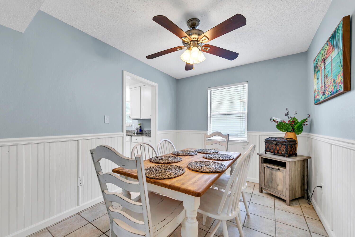 Un coin repas avec une table en bois, des chaises blanches, un ventilateur de plafond et une fenêtre avec des stores.