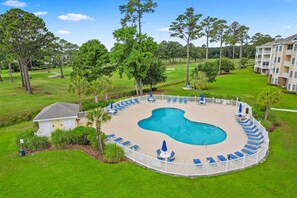 A circular pool with lounge chairs and umbrellas, surrounded by a white fence, set in a grassy area with trees and a building in the background.