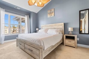 A bedroom with a large bed, a nightstand, a bedside table, a wall-mounted mirror, and a window with curtains.