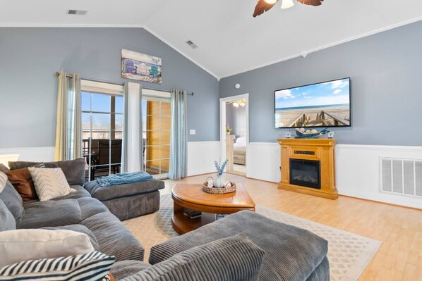 A living room with a grey sofa set, a wooden coffee table, a fireplace, and a large TV mounted on the wall.