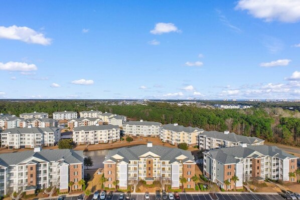 A residential complex with multiple apartment buildings, a parking area, and a view of a forested area in the background.