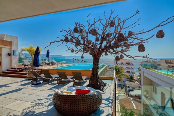 A rooftop terrace with a decorative tree sculpture, lounge chairs, a pool, and a view of the sea.