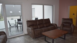 A living room with a brown leather sofa, a wooden coffee table, and a brown leather armchair. There is a sliding glass door leading to an outdoor area with a table and chairs. - Hunsr./Reich vacation apartment in the countryside suitable for family a. fitter (Reich)