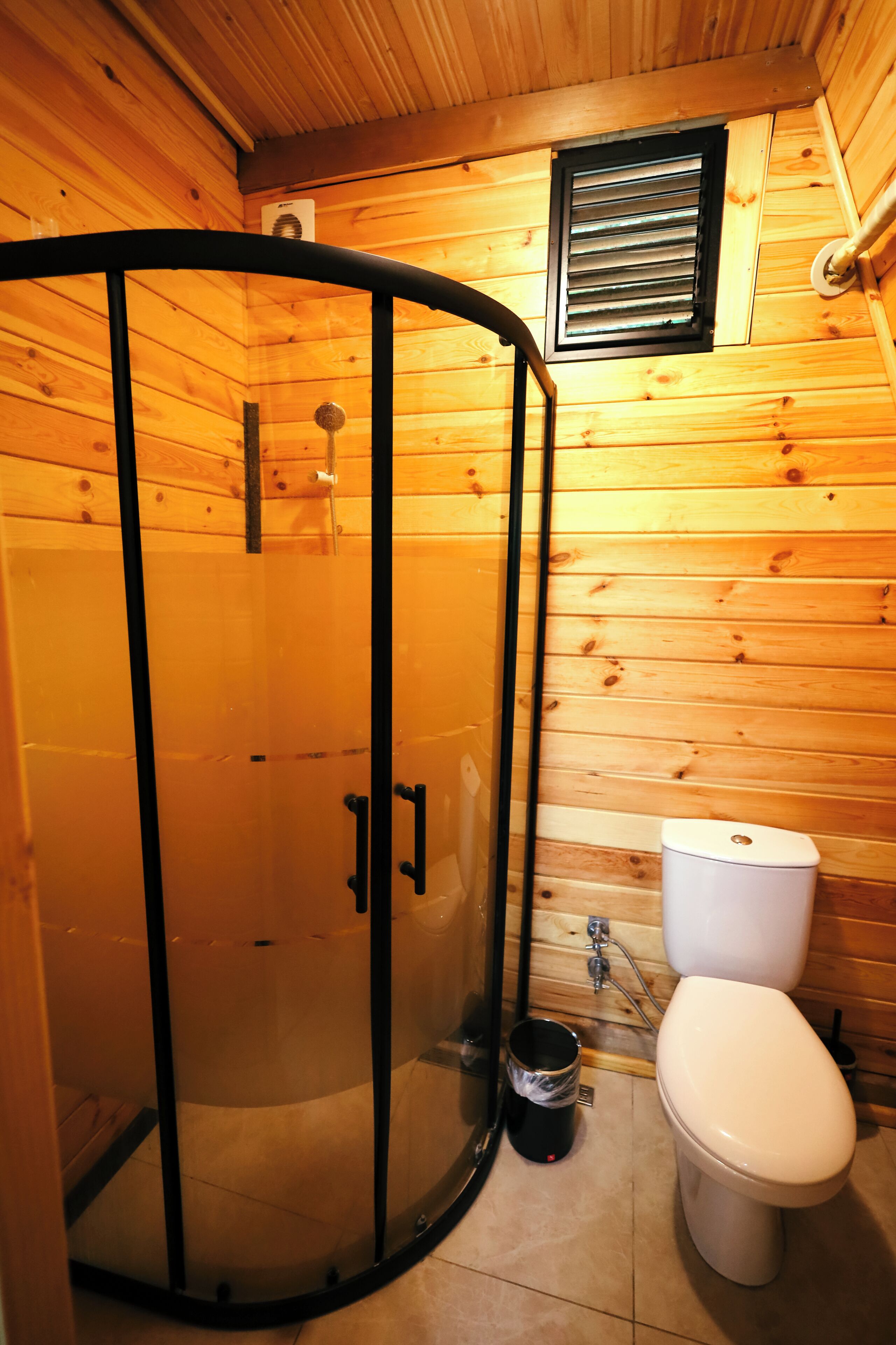 A wooden cabin bathroom with a glass-enclosed shower, a toilet, and a window.