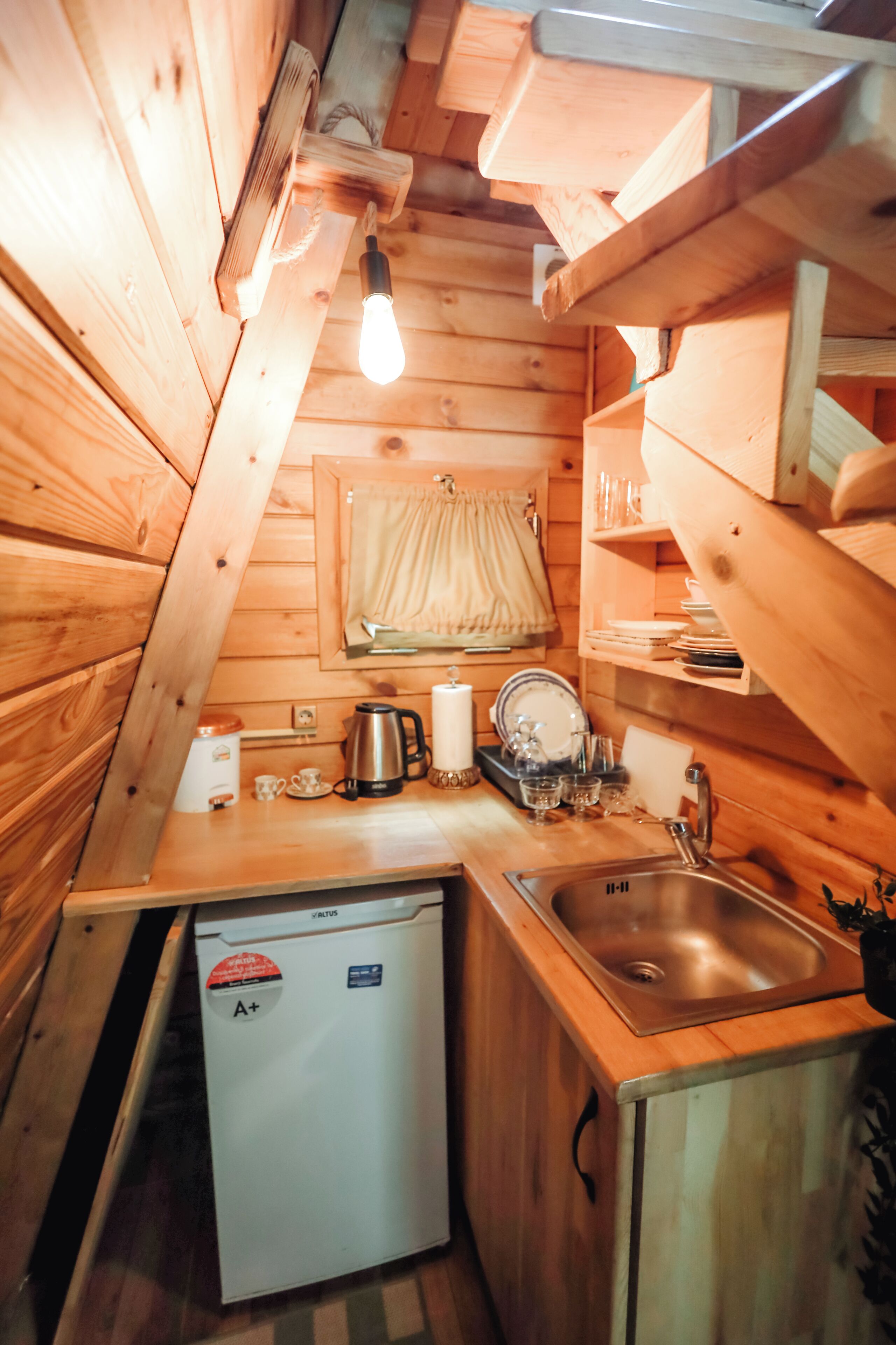 A compact wooden cabin kitchen with a sink, a small refrigerator, and a light bulb hanging from the ceiling.