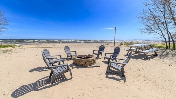 A beach setting with Adirondack chairs arranged around a stone fire pit, overlooking the ocean. - State Ave|Beach|King Beds|Central AC (Alpena)